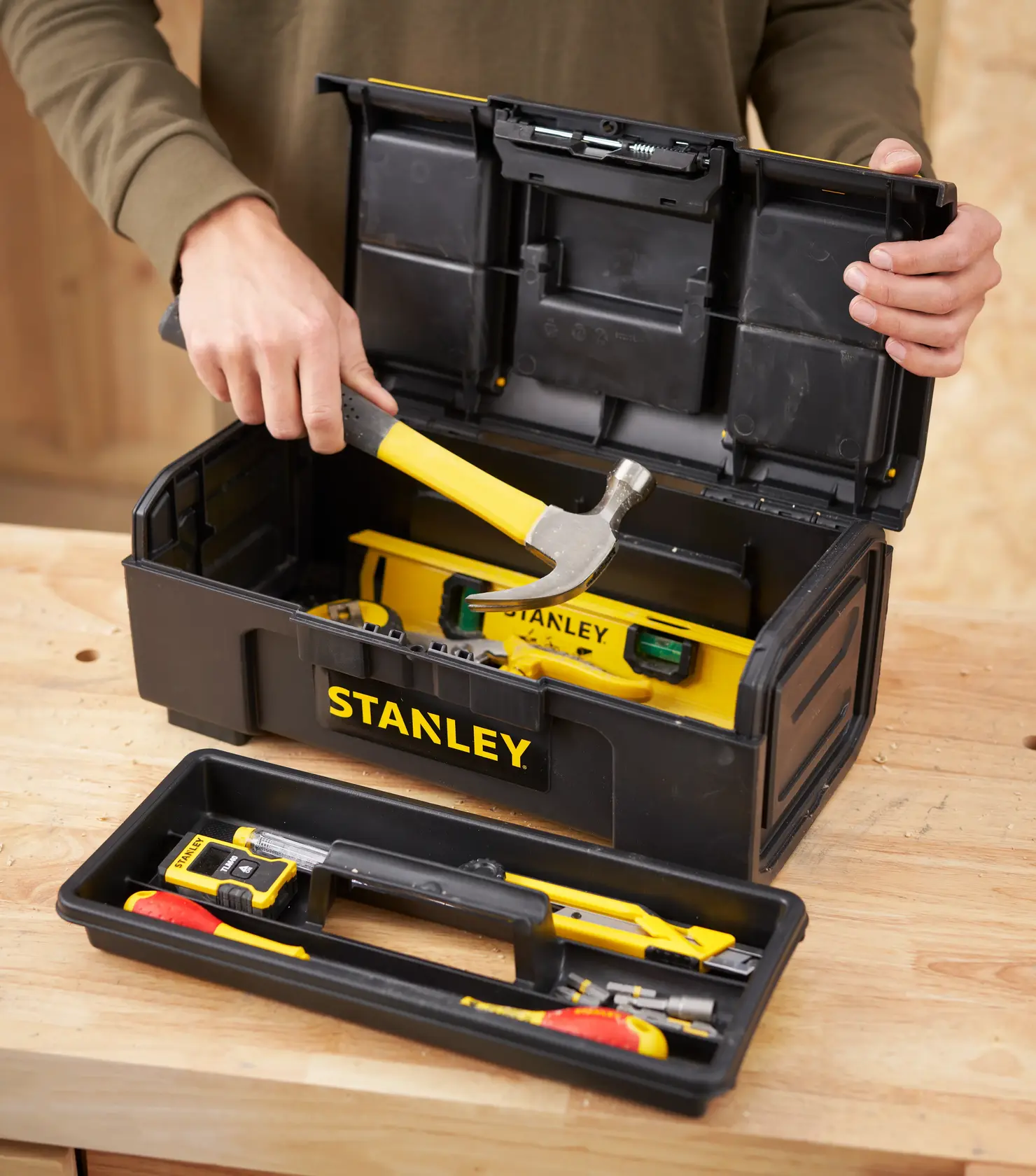 Photo of STANLEY 1-79-216 toolbox with yellow hand tools, open on a wooden table, person holding hammer.