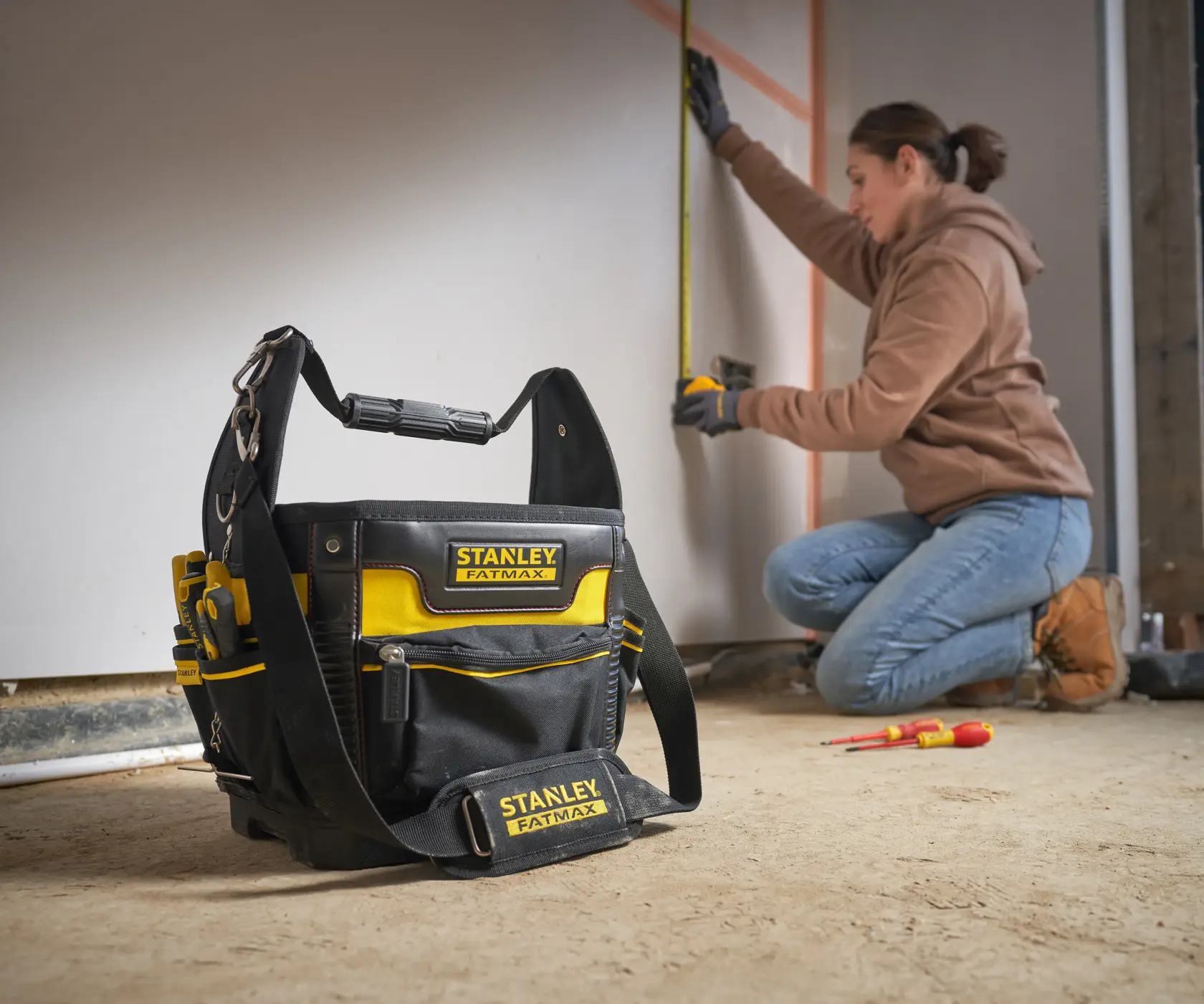 STANLEY 1-93-952 tool bag with yellow and black design on floor, worker measuring wall in background.