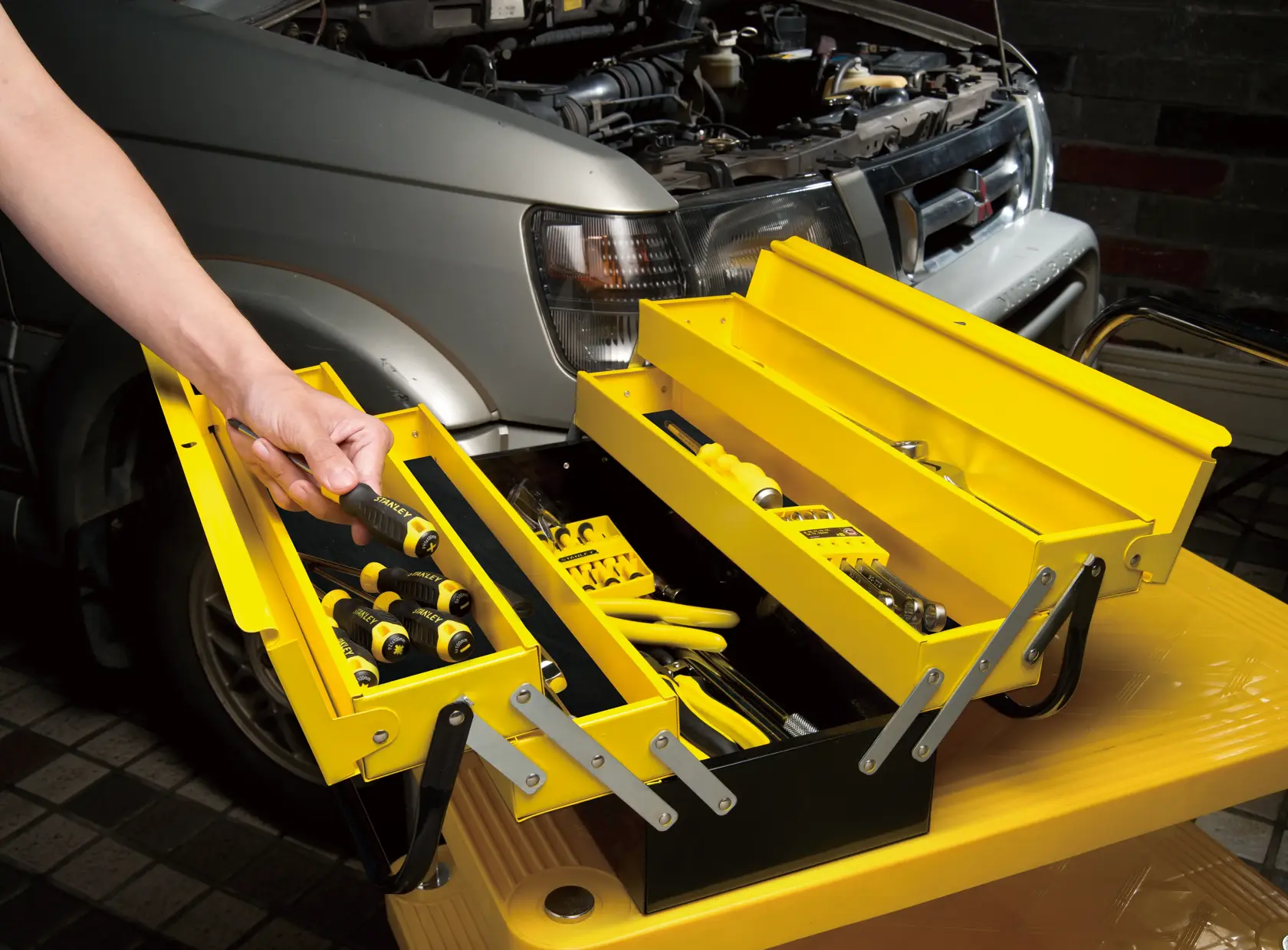 STANLEY 1-94-738 yellow metal tool box with hand tools, photography side angle, used near car engine.