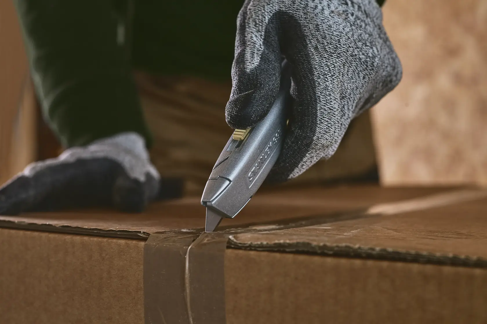 STANLEY 2-10-099 utility knife in gloved hand cutting tape on a cardboard box, close-up action shot.