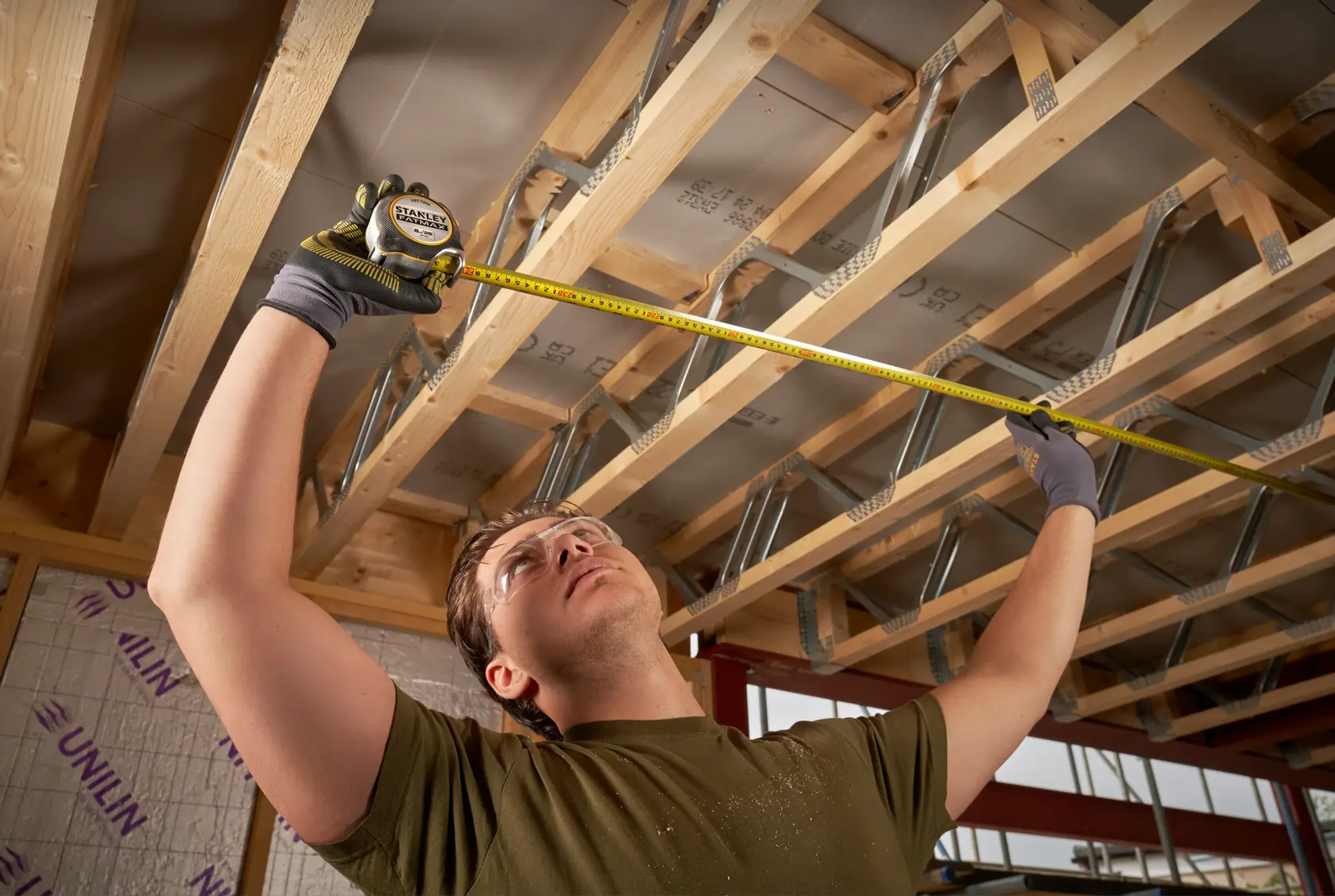 STANLEY FMHT38218-5 tape measure used by person wearing gloves to measure wooden ceiling beams in construction site.