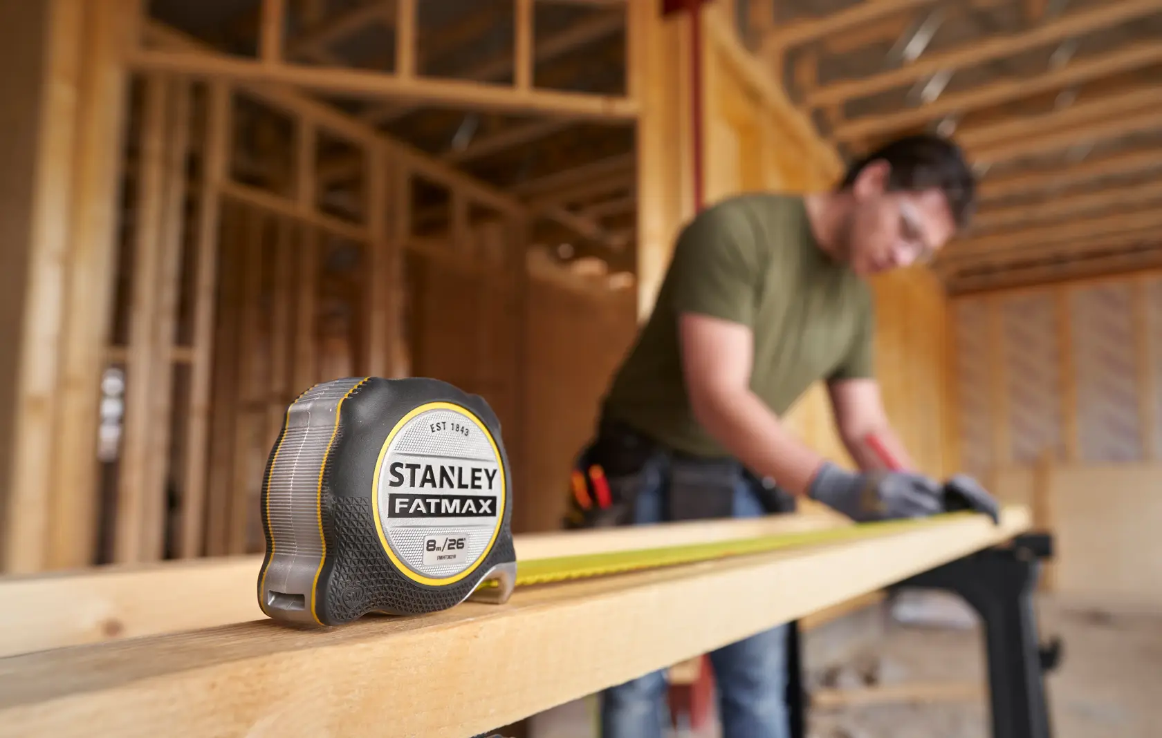 Photo of a STANLEY FATMAX tape measure, SKU FMHT38218-5, on a wooden beam at a construction site, with a worker in the background.