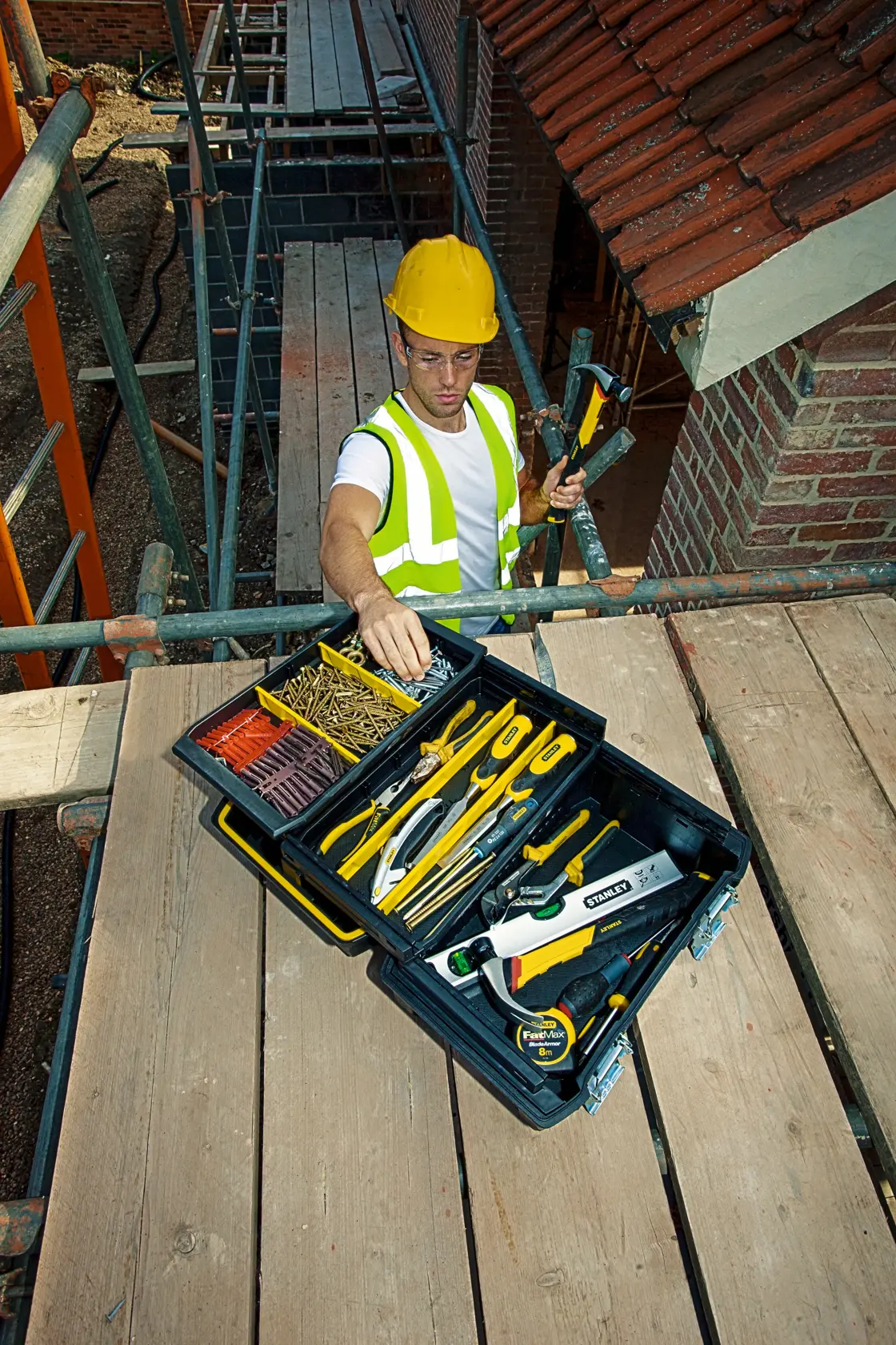 Photo of STANLEY FMST1-71219 toolbox with assorted tools on scaffolding, worker in safety gear nearby.