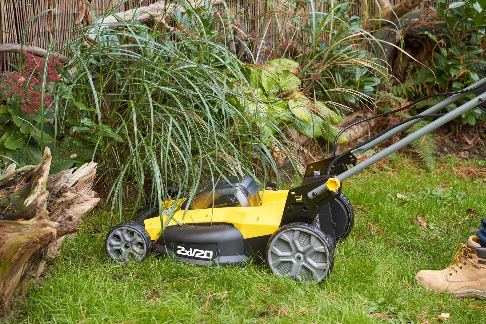 STANLEY SFMCMWS251B yellow cordless lawn mower in use cutting grass near plants outdoors.