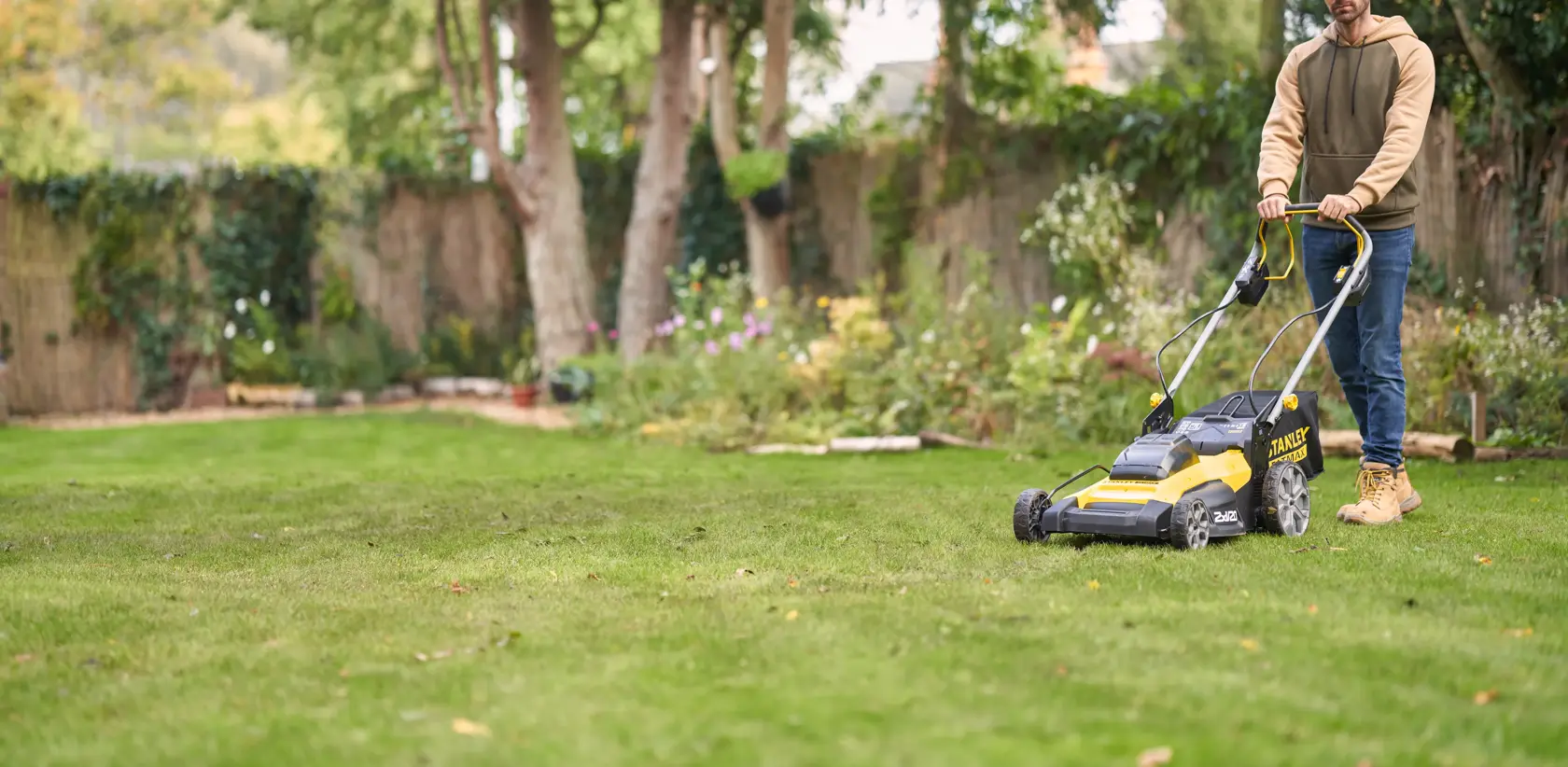 Photo of STANLEY SFMCMWS251B cordless lawn mower in use on a green lawn, garden and trees in background.