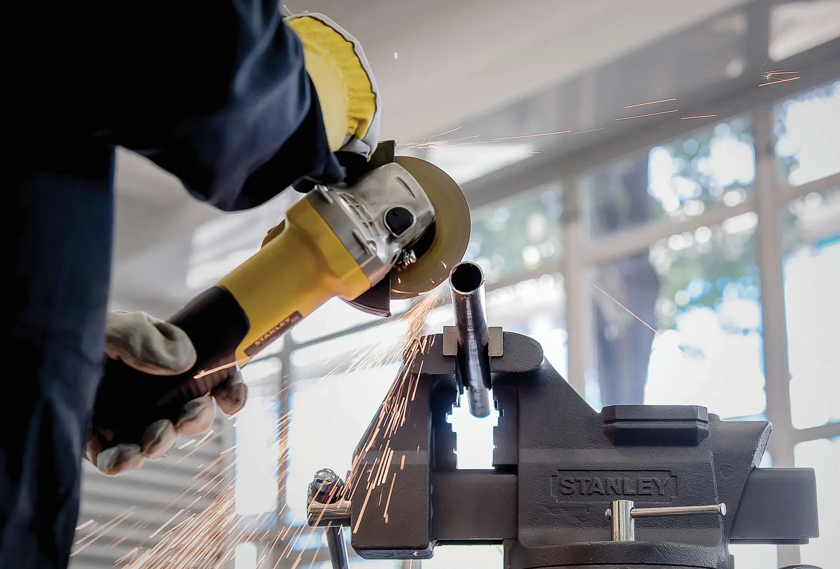STANLEY STA8061 bench vise holding metal pipe, with sparks flying as worker uses angle grinder.