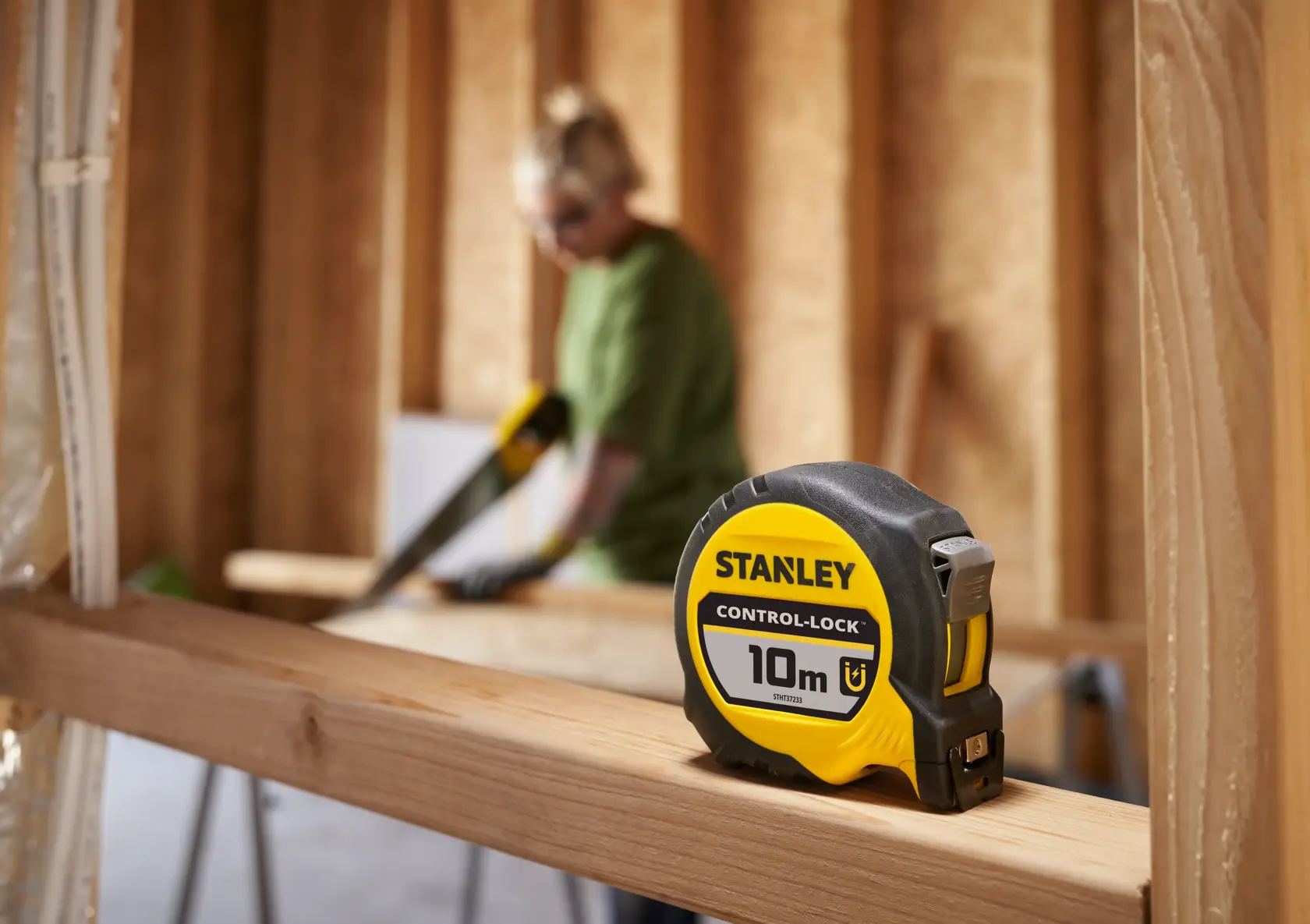 Stanley STHT37233-0 tape measure, 10m, yellow and black, on wood beam with blurred workspace background.