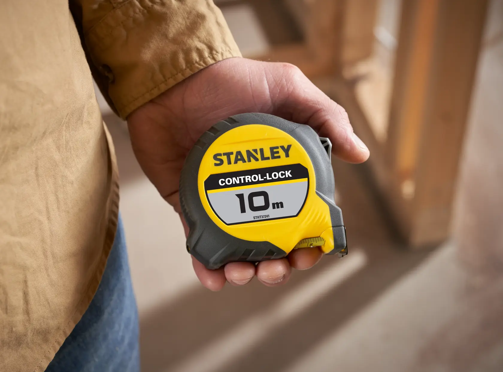 Hand holding a STANLEY Control-Lock 10m tape measure with yellow and gray casing. Indoor construction setting.