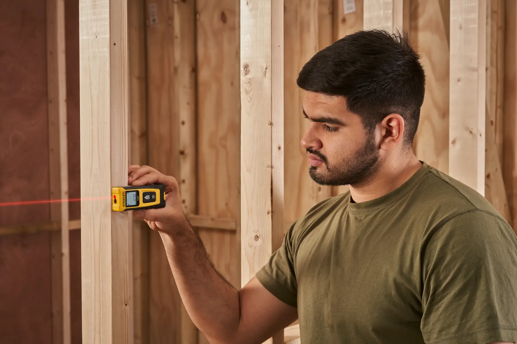Photo of STANLEY STHT77065-0 laser distance measurer in use, measuring wood studs in a construction setting.