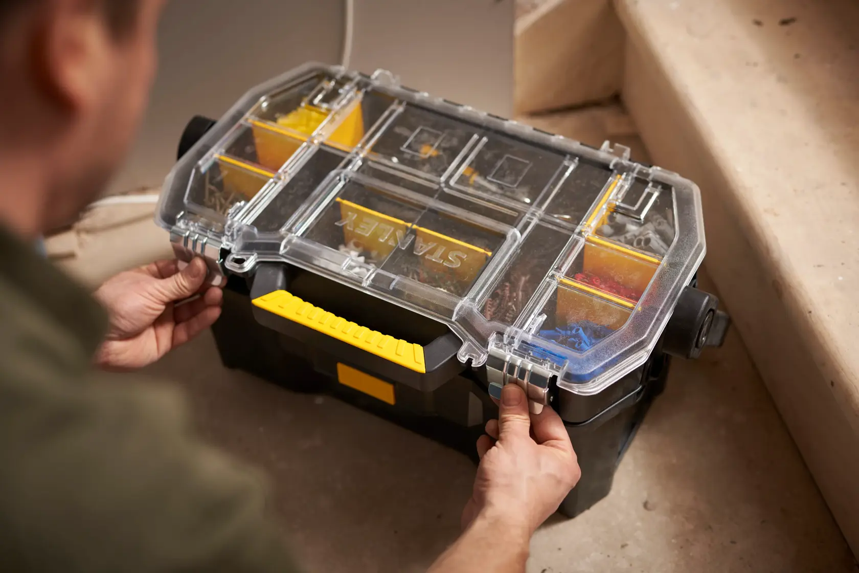 STANLEY STST1-70317 tool organizer with clear lid and yellow handle, being opened by hands on a work site.